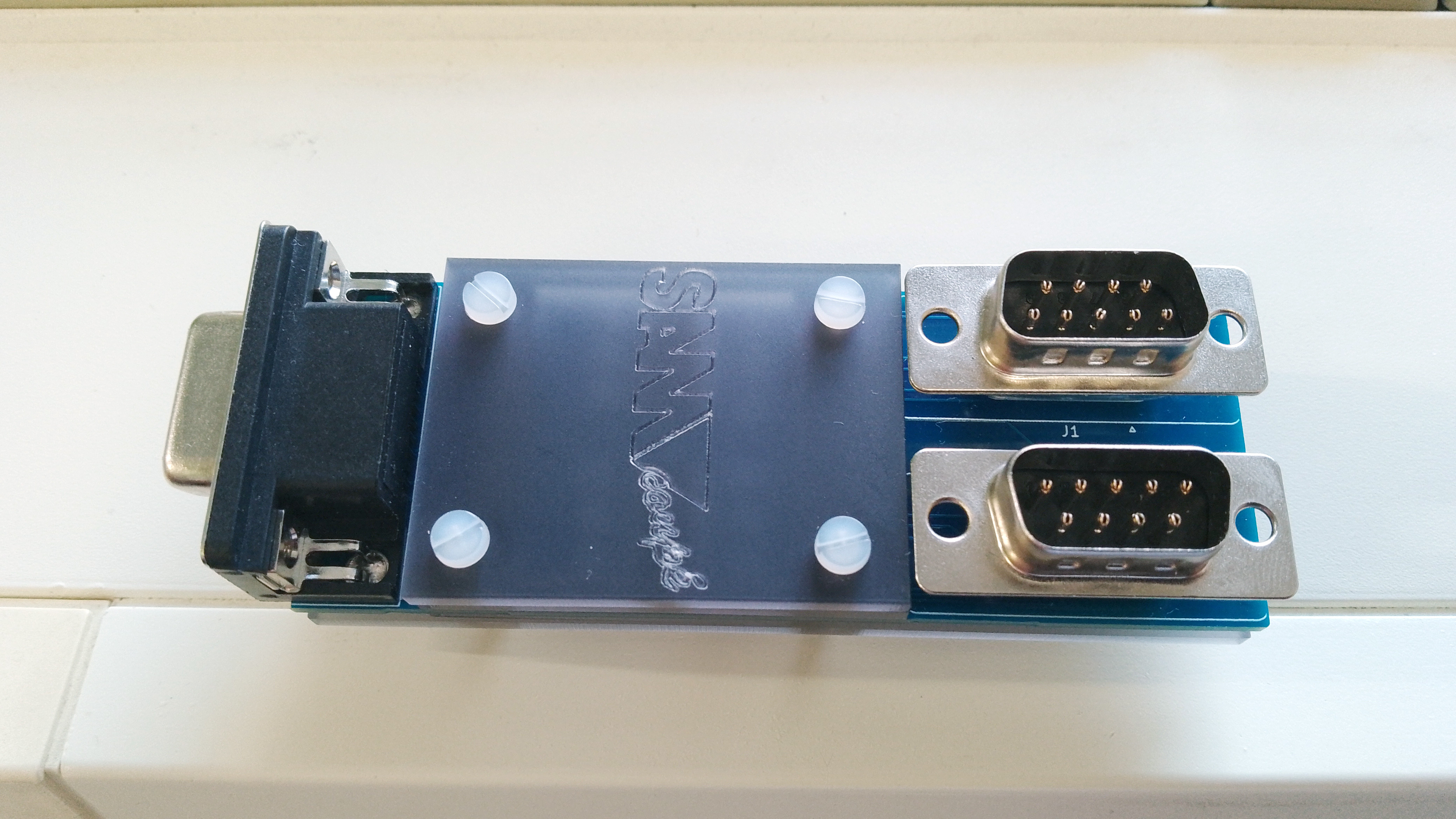 Small PCB, partially enclosed in plastic, with a single female 9-pin D-Sub connector at one end and two male 9-pin D-Sub connectors at the other. The plastic casing has the SAM Coupé logo etched on the top.